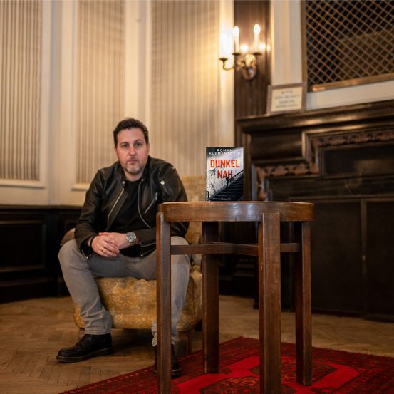 Ein Mann sitzt auf einem Stuhl in einem Raum, hält ein Buch mit dem Titel Dunkel Nah und trägt eine Lederjacke und eine Uhr.