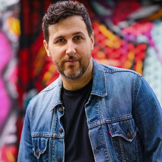 Ein Mann mit Bart und Schnurrbart lächelt vor einer farbenfrohen Graffiti-Wand und trägt eine Jeansjacke.