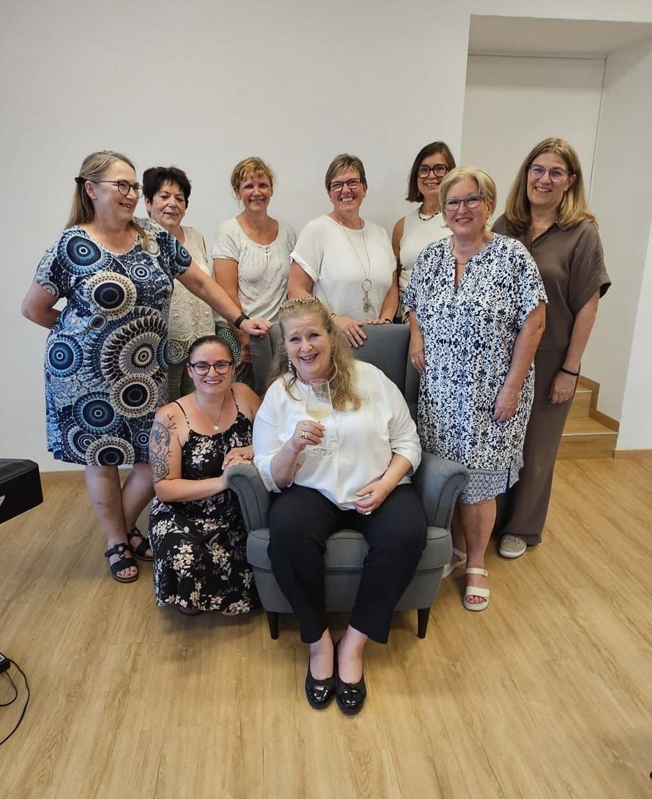 Eine Gruppe von Frauen posiert für ein Foto in einem Raum mit Holzböden. Eine Frau im Stuhl hält ein Weinglas.