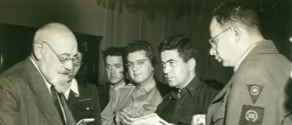 Ein Schwarz-Weiß-Foto zeigt eine Gruppe von Männern in Uniformen und Anzügen. Der Mann in der Mitte hält ein Stück Papier, während ein anderer Mann im Anzug zu ihm spricht.