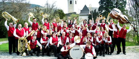 Eine Band mit roten Westen posiert vor einer Kirche. Sie halten verschiedene Instrumente wie Trompeten, Posaunen und Trommeln.