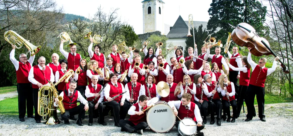 Eine Band mit roten Westen posiert vor einer Kirche. Sie halten verschiedene Instrumente wie Trompeten, Posaunen und Trommeln.