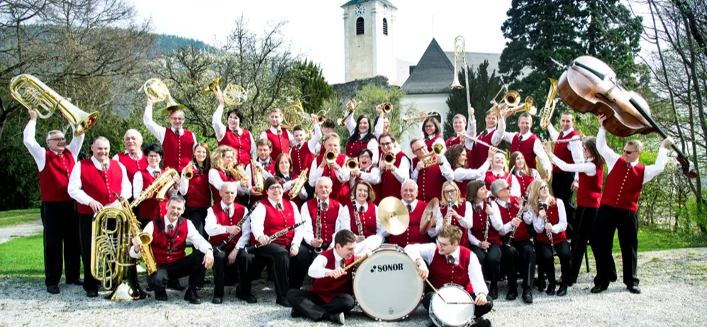 Eine große Gruppe von Musikern in roten und weißen Westen posiert mit ihren Instrumenten vor einer Kirche.