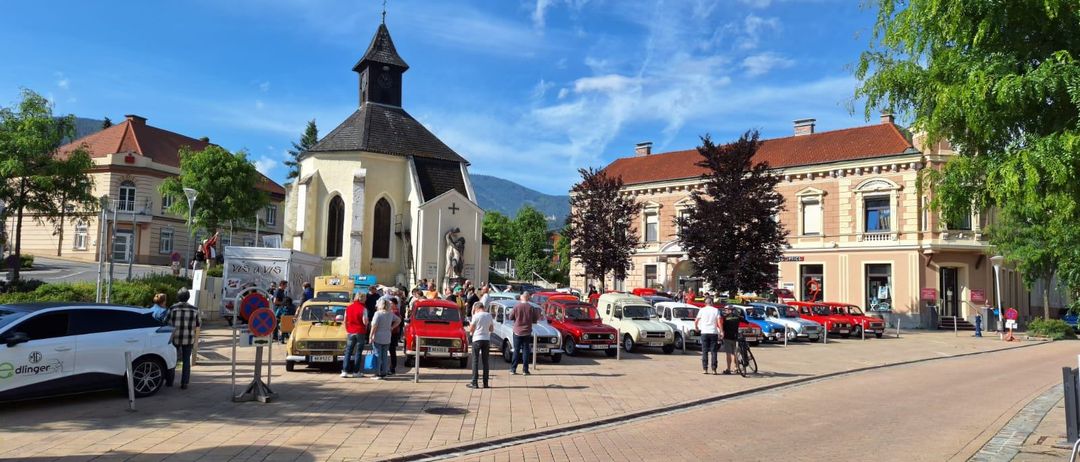 Ein Stadtplatz mit vielen geparkten Autos, darunter ein gelbes und ein rotes. Eine Kirche steht im Hintergrund mit einer Statue auf der Vorderseite.