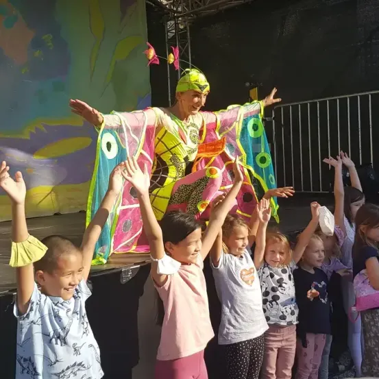 Eine farbenfrohe Darstellerin unterhält eine Gruppe von Kindern auf der Bühne, alle heben ihre Hände vor Freude in die Höhe.
