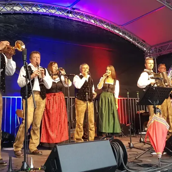 Eine Gruppe von Musikern tritt auf der Bühne auf, darunter Trompete, Klarinette, Akkordeon und Gesang. Sie sind in traditioneller Kleidung gekleidet, mit Scheinwerfern und Mikrofonen vor ihnen.