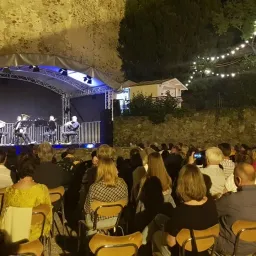 Eine Gruppe Musiker tritt nachts auf einer Bühne auf. Das Publikum, das in Stühlen sitzt, beobachtet sie unter einem Vordach mit Lichterketten.