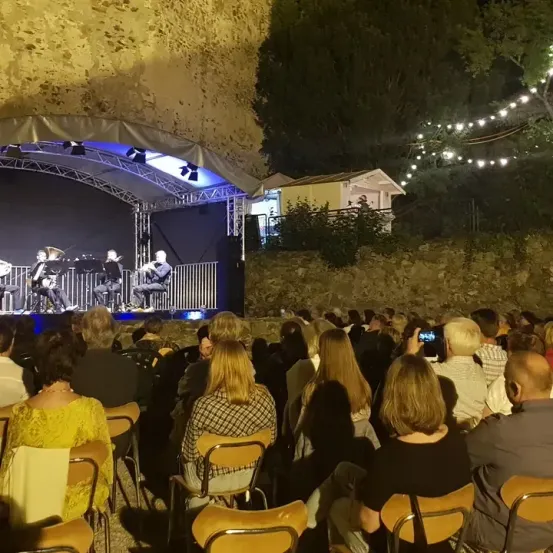 Eine Gruppe Musiker tritt nachts auf einer Bühne auf. Das Publikum, das in Stühlen sitzt, beobachtet sie unter einem Vordach mit Lichterketten.