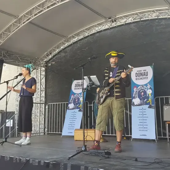 Eine Frau und ein Mann treten auf der Bühne auf. Der Mann spielt Gitarre und die Frau singt. Hinter ihnen stehen zwei Plakate.