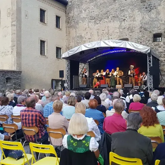 Eine Gruppe von Musikern tritt auf einer Bühne vor einem großen Publikum auf. Die Bühne ist mit einem Zelt, Lautsprechern und Beleuchtung ausgestattet. Das Publikum sitzt auf Stühlen und beobachtet aufmerksam die Aufführung.