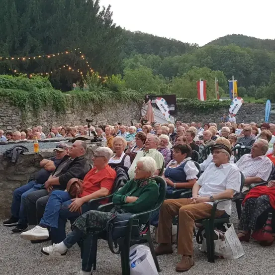 Eine große Gruppe von Menschen sitzt in Stühlen in einem Außenbereich. Sie schauen sich eine Veranstaltung an. Die Menschen tragen Brillen, Hüte und Schuhe. Einige von ihnen haben Handtaschen dabei. Es gibt einen verschwommenen Blick auf eine Wand mit einem Banner und Flaggen. In der Ferne sind Bäume und Berge zu sehen.