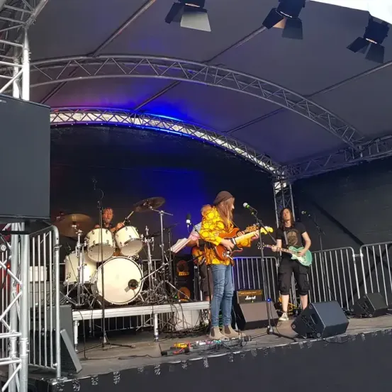 Eine Band spielt auf der Bühne mit einem Schlagzeuger, einem Gitarristen und einem Sänger, unter Scheinwerfern und mit Lautsprechern auf beiden Seiten.