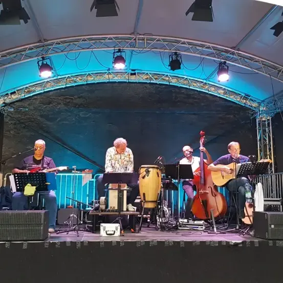 Eine Band mit fünf Männern spielt auf der Bühne unter einem Metallgerüst mit Lichtern. Jeder Mann spielt ein anderes Instrument. Die Bühne ist mit Musikständern und Lautsprechern ausgestattet.