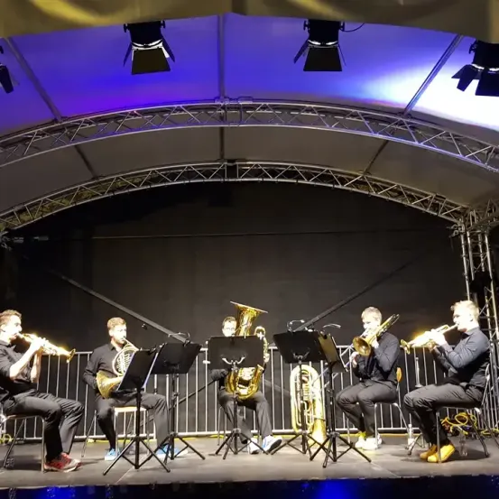 Ein Blechbläserensemble aus sechs Musikern tritt auf der Bühne unter blauem Licht auf. Sie sitzen auf Stühlen und spielen verschiedene Blechblasinstrumente. Die Bühne hat einen Metallrahmen und einen schwarzen Hintergrund.