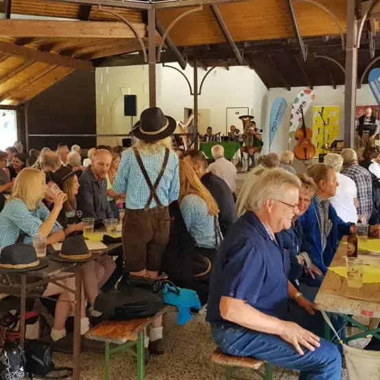 In einem geschäftigen Veranstaltungsort sitzen eine Gruppe von Menschen um Tische herum. Eine Frau mit Hut und Hosenträger steht. Hinter ihnen spielt eine Band auf der Bühne. Die Umgebung ist lebhaft, mit verschiedenen Getränken und Gegenständen auf den Tischen.