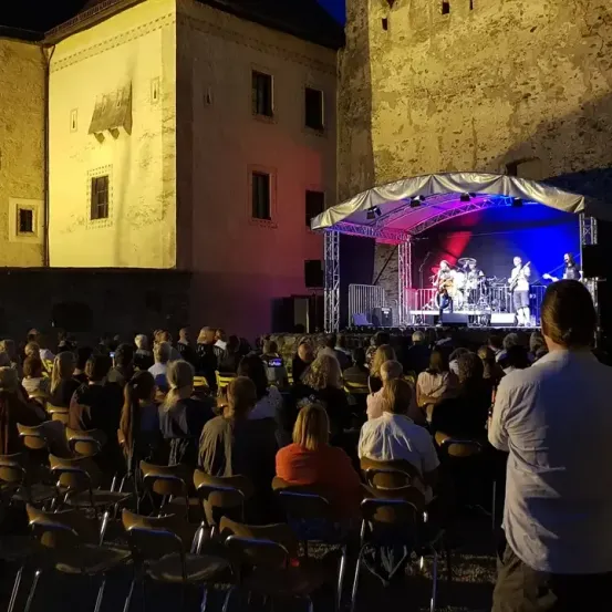 Ein nächtliches Konzert mit einer Band auf der Bühne, beleuchtet von Lichtern, vor einem Publikum, das in Stühlen sitzt.