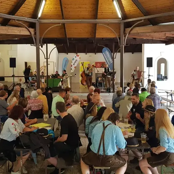 Innenveranstaltung mit Bühnenshow, Leute an Tischen sitzend, zwei Musiker spielen Blechblasinstrumente, einer mit Cello, Bannerwerbung an Ständern und hölzerne Decke.