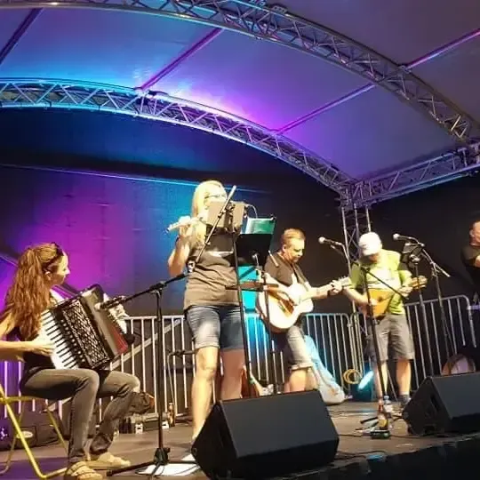 Eine Gruppe von Musikern tritt auf einer Bühne auf. Eine Frau spielt Flöte, ein Mann Gitarre. Ein anderer Mann spielt Banjo, ein anderer Akkordeon. Die Bühne hat Metallgeländer und Lautsprecher.