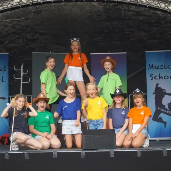Eine Gruppe junger Mädchen in verschiedenen farbigen T-Shirts tritt auf der Bühne auf, lächelt und posiert für die Kamera. Die Bühne hat einen Lautsprecher und einen Hintergrund mit dem Text 'Musikschule'.