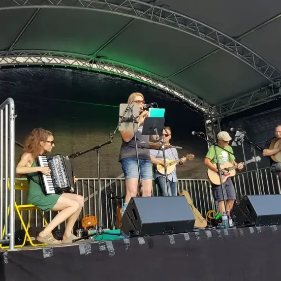 Eine Gruppe von Musikern tritt auf einer Bühne unter einem Vordach auf. Eine Frau spielt Akkordeon, während ein Mann in ein Mikrofon singt. Ein anderer Mann spielt Gitarre und ein Mann dahinter spielt Banjo. Lautsprecher stehen auf der Bühne.