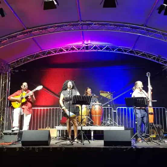 Eine Band tritt auf der Bühne auf. Eine Frau in der Mitte singt, während andere Instrumente wie Schlagzeug, Gitarre und Cello spielen. Scheinwerfer sind an der Decke montiert.