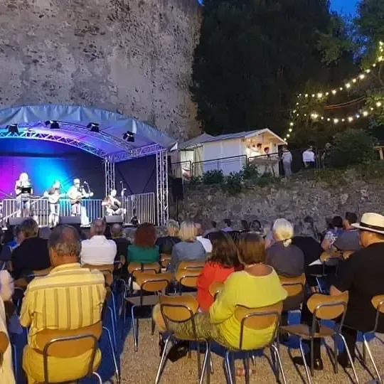 Eine Gruppe von Menschen besucht ein nächtliches Open-Air-Konzert mit Bühne, Beleuchtung und einem Hintergrund aus Bäumen und Felsen. Die Künstler spielen Gitarren und andere Instrumente.