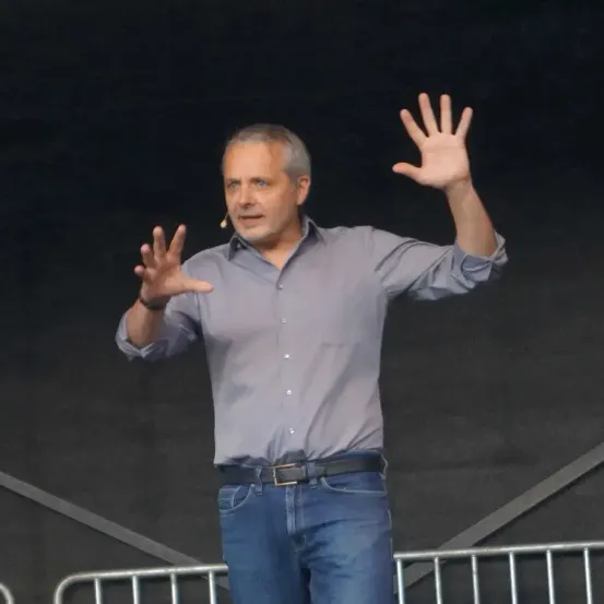 Ein Mann steht auf der Bühne, trägt ein graues Hemd und blaue Jeans. Er hat die Hände erhoben und spricht zu einem Publikum.
