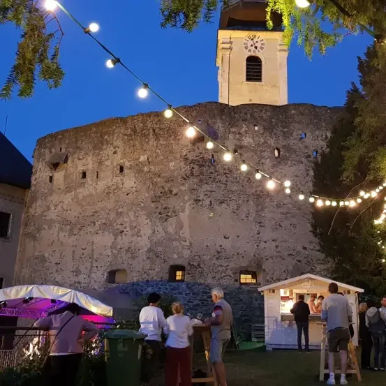 Eine Gruppe von Menschen versammelt sich bei Nacht vor einer alten Burg mit einem Glockenturm und Lichterketten. Ein Imbissstand ist aufgestellt, an dem mehrere Personen stehen.