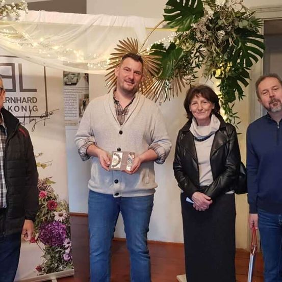 Vier Menschen stehen lächelnd in einem Raum mit Dekoration. Ein Mann in der Mitte hält eine Auszeichnung. Hinter ihnen ist eine Blumendekoration und ein Banner.