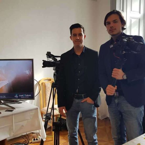 Zwei Männer stehen neben einer Kamera-Einrichtung in einem Raum. Ein Mann hält eine Kamera, während der andere neben ihm steht. Hinter ihnen ist ein Fernsehbildschirm, der ein Hochzeitsvideo zeigt.