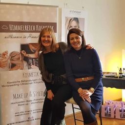 Zwei Frauen stehen vor einem Banner, das für 'Himmelreich Kosmetik' wirbt. Sie lächeln und posieren für ein Foto. Dahinter steht ein Schreibtisch mit Kosmetikprodukten. Das Banner zeigt das gestylte Haar einer Frau und weitere angebotene Schönheitsdienstleistungen.