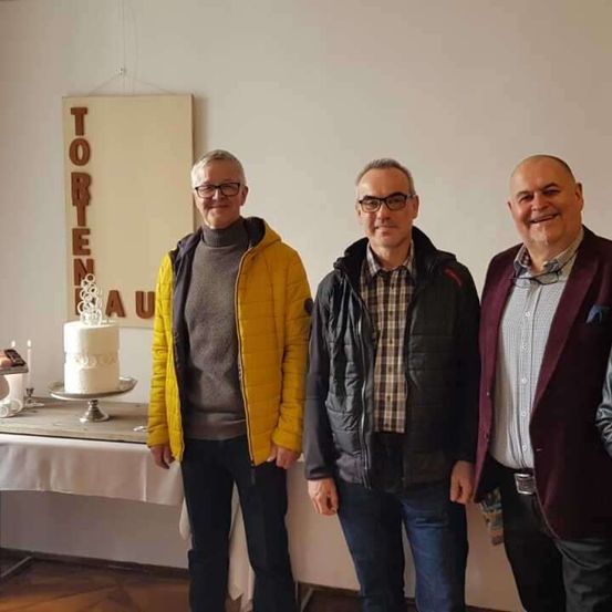 Vier Männer stehen zusammen vor einer Wand mit dem Wort 'Tortenhaus'. Hinter ihnen steht ein Tisch mit einem Kuchen, und sie alle lächeln.