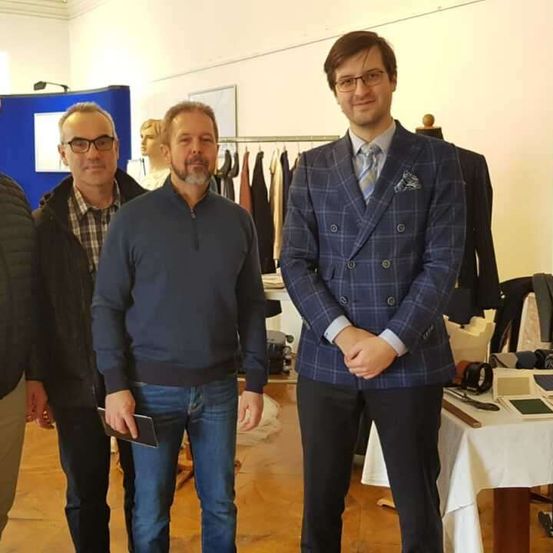 Vier Männer stehen in einem Raum mit Kleidern an Kleiderbügeln hinter ihnen, einer hält ein Buch, die anderen tragen Brillen und Anzüge.