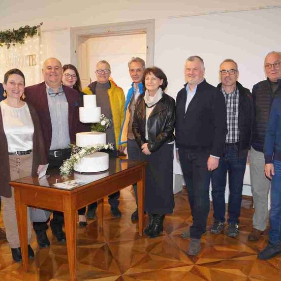 Eine Gruppe von Menschen steht um einen Tisch mit einer Torte und lächelt für ein Foto. Die Torte ist mit Blumen verziert. Der Raum hat weiße Wände und einen gemusterten Boden.