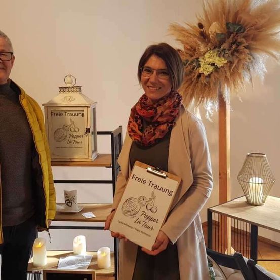 Ein Mann und eine Frau stehen nebeneinander und lächeln beide. Die Frau hält eine Mappe mit einem Buch mit dem Titel 'Freie Trauung'. Dahinter steht ein Regal mit einer Laterne und Kerzen.