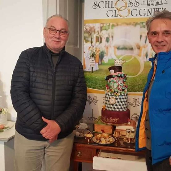 Zwei Männer stehen neben einem Tisch mit Desserts vor einem Banner für Schloss Gloggnitz.