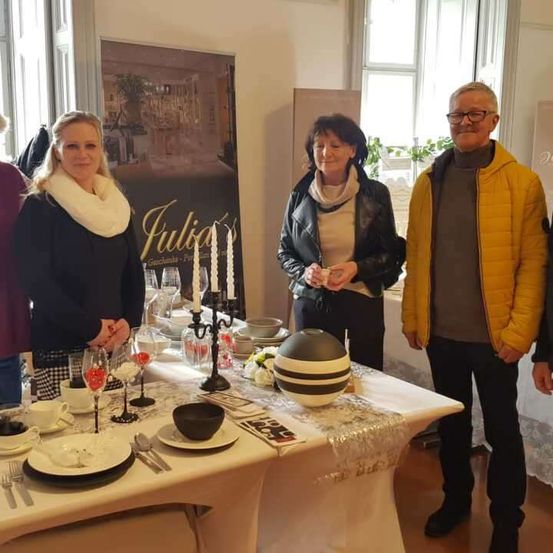 Vier Personen stehen um einen Tisch herum, möglicherweise bei einer Veranstaltung. Der Tisch ist mit Tellern, Schüsseln, Weingläsern und Besteck gedeckt. Eine Frau auf der linken Seite lächelt, während eine andere Frau in der Mitte einen Becher hält. Ein Mann auf der rechten Seite trägt eine Brille und eine gelbe Jacke. Hinter ihnen befindet sich ein Banner mit dem Wort 'Julia's'.