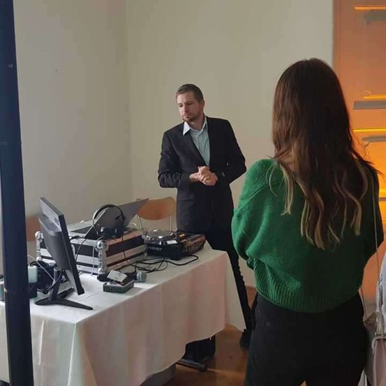 Ein Mann im Anzug steht hinter einem Schreibtisch mit Laptop, Kopfhörern und DJ-Mixer und spricht mit einer Frau in einem grünen Pullover.