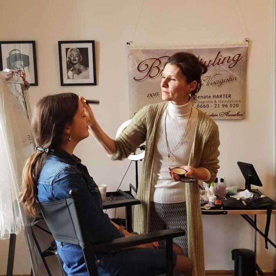 Eine Frau schminkt eine andere Frau, die in einem Stuhl in einem Salon sitzt. Ein Schild im Hintergrund lautet 'Schönheit'.