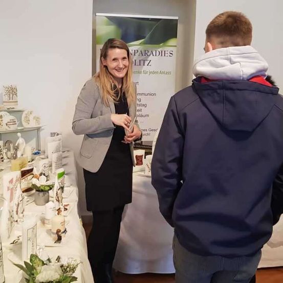 Eine Frau steht lächelnd hinter einem Tisch mit verschiedenen Gegenständen. Ein Junge steht vor ihr. Hinter ihnen befindet sich ein Banner mit Text.