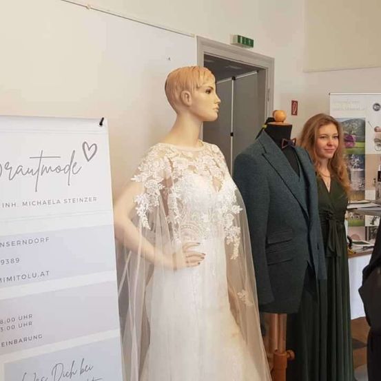 Eine Puppe in einem Hochzeitskleid steht vor einem weißen Banner mit Brautmode drauf. Eine Frau steht neben einer Puppe in einem Anzug.