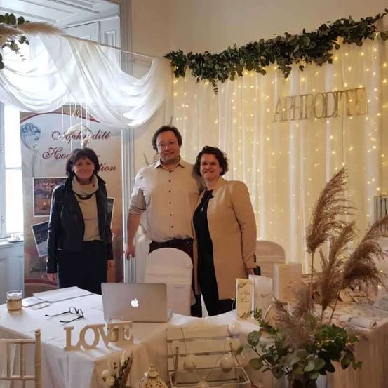 Drei Personen stehen an einem Schreibtisch in einem Raum mit weißem Hintergrund, Lichterketten und dem Wort Aphrodite. Der Tisch hat einen Laptop, ein Schild mit der Aufschrift LOVE und andere Gegenstände.