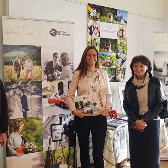 Zwei Frauen stehen neben einer Wand mit Hochzeitsfotos. Eine hält ein Büchlein und lächelt. Die andere hält ein Buch. Ein Tisch mit Gegenständen steht hinter ihnen.