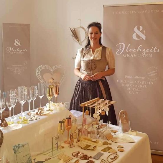 Eine Frau steht hinter einem Tisch mit verschiedenen Artikeln zum Verkauf, einschließlich Weingläser, Metalldekorationen und kleine Objekte, in einem Raum mit einer weißen Wand und einem großen Schild mit Text in Deutsch.