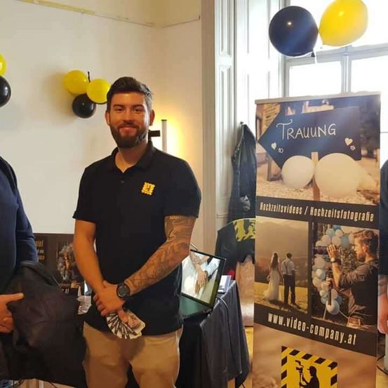 Ein Mann mit einer Tätowierung am Arm steht neben einem Banner, das Hochzeitsvideos und -fotos bewirbt. Er hält einen Stapel Geld. Hinter ihm hält eine andere Person eine Tasche. Ein Tisch mit einem Laptop ist in der Nähe, und Luftballons hängen an der Wand.