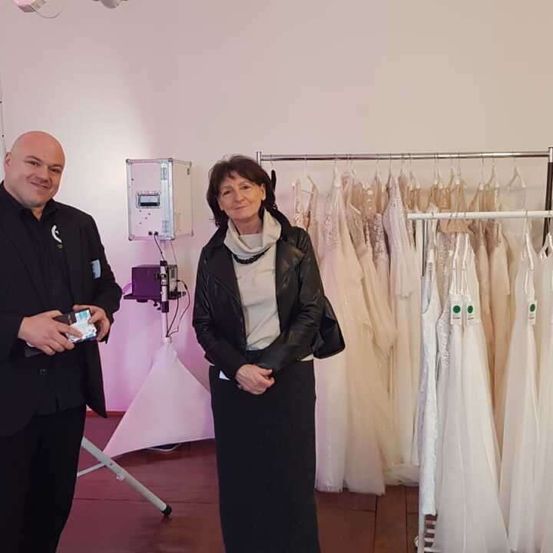 Ein Mann und eine Frau stehen nebeneinander vor einem Ständer mit Brautkleidern. Der Mann hält eine kleine Schachtel und lächelt. Die Frau trägt eine schwarze Lederjacke und steht neben einem Ständer mit weißen Brautkleidern. Dahinter steht ein weißes Zelt.