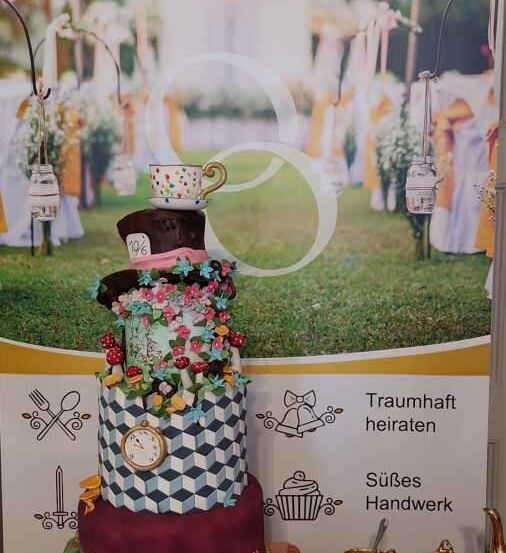 Ein großer mehrstöckiger Kuchen mit Mad Hatter-Thema auf einem Tisch mit verschiedenen Dekorationsartikeln, vor einem Hintergrund mit einem Grasfeld und weißen Zelten.