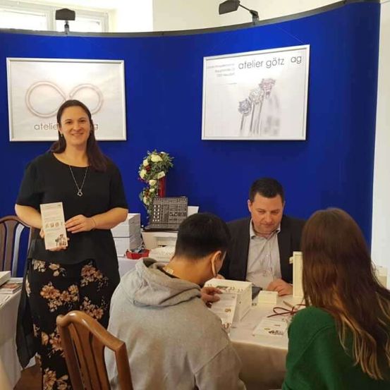 Eine Frau in einem schwarzen Kleid hält ein Buch in der Hand und steht vor einer blauen Wand. Hinter ihr sitzt ein Mann an einem Tisch mit einer Blumenarrangement und anderen Gegenständen. Andere Personen sitzen um den Tisch herum, und eine schaut in ein Buch.