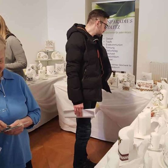Ein Mann in einer schwarzen Jacke schaut sich Gegenstände auf einem Tisch an, neben ihm steht eine Frau in einem blauen Shirt. Sie sind bei einer Veranstaltung, mit einer weißen Tischdecke und einer Ausstellung von Gegenständen hinter ihnen.