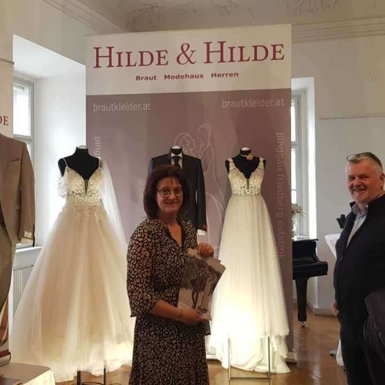 Eine Frau hält ein Buch vor einer Brautkleid-Ausstellung, neben ihr steht ein lächelnder Mann. Hinter ihnen sind Hochzeitskleider auf Mannequins und ein Banner mit der Aufschrift 'HIlDE & HIlDE' zu sehen.